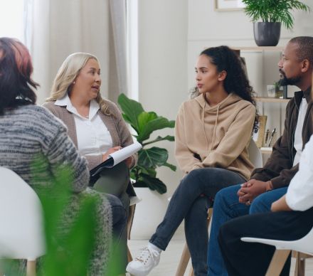 Female counselor with clipboard leading a small group therapy session indoors.
