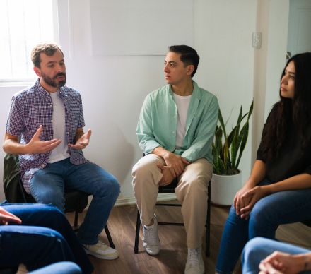 Man speaking and gesturing during a small group therapy session in a bright room.