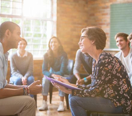 Therapist with clipboard engaging one-on-one with a patient during a group therapy session.