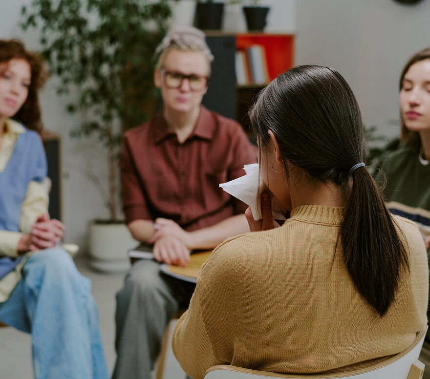 Diverse group of people in a support circle, woman in foreground with tissue, compassionate peer support setting