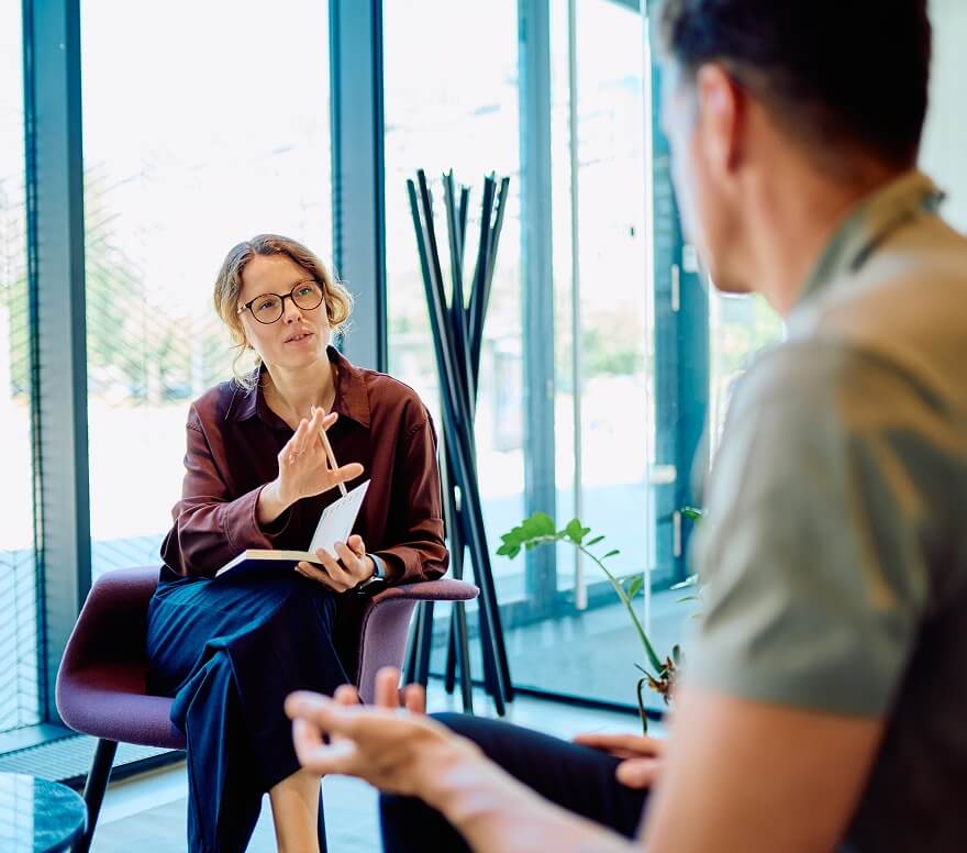 Female addiction counselor consulting with male patient in bright office setting with plants and natural light
