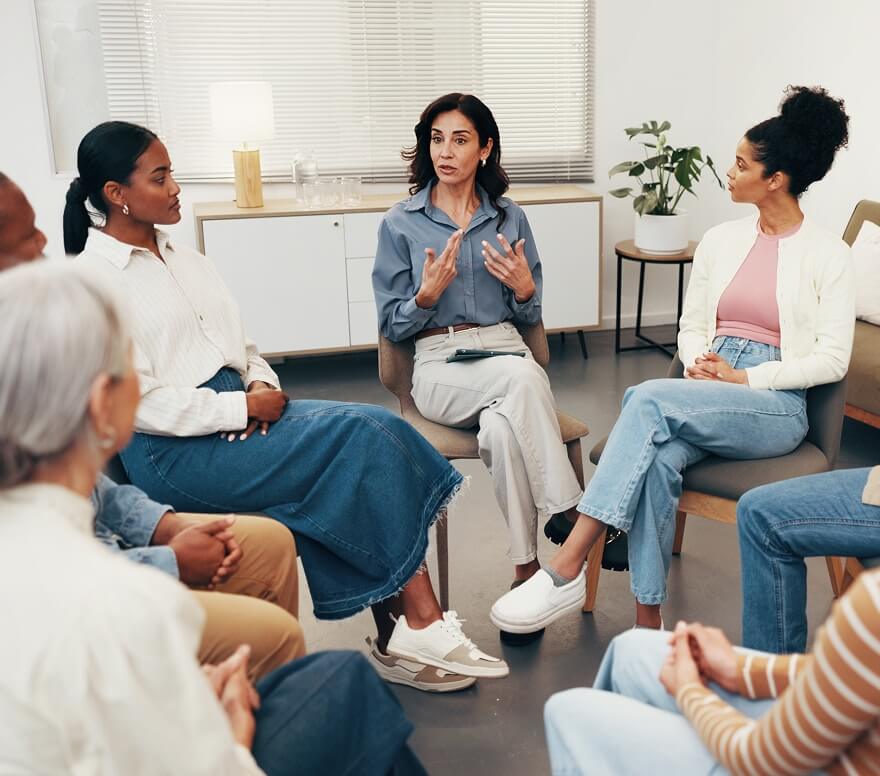 peer-centers-and-recovery-housing Woman leading a group therapy discussion in a circle of chairs.