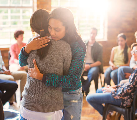 Two women embracing during a group therapy session, with other group members seated in a circle.