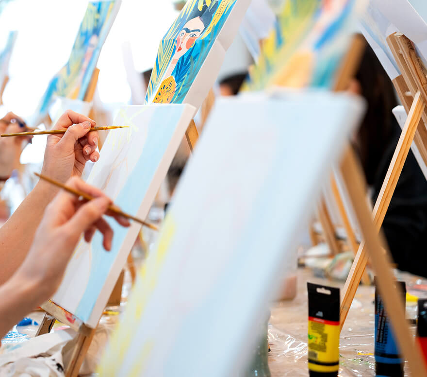 A group painting workshop with participants holding brushes and working on canvases displayed on easels